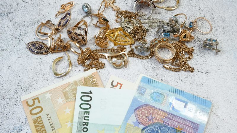 Sammlung von goldenen und silbernen Schmuckstücken – darunter Ringe, Halsketten und Ohrringe – auf einer hellen, strukturierten Oberfläche verstreut. Im Vordergrund liegen mehrere Euro-Banknoten (50, 100 und 200 Euro), was auf einen Kontext von Reichtum, Handel oder Bewertung hinweist.